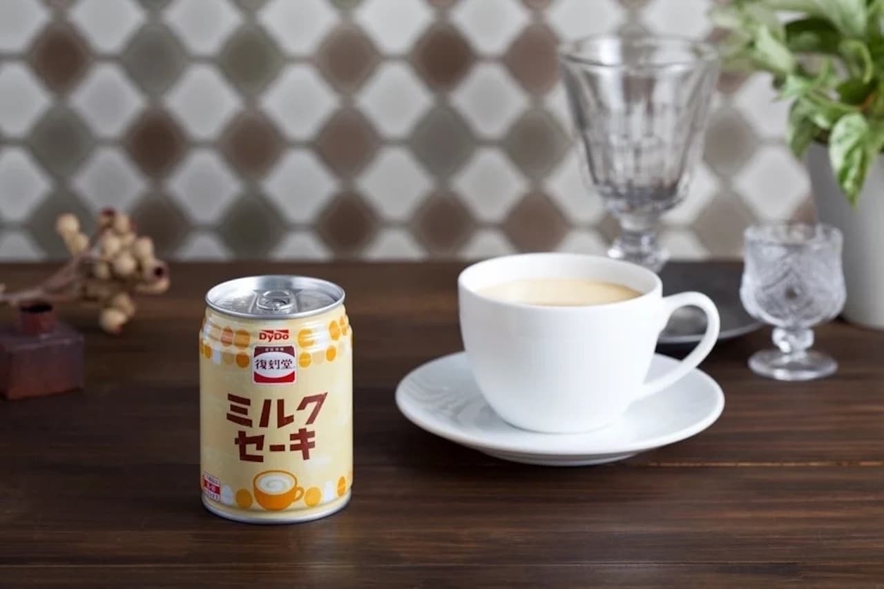 A product photo of a DyDo can labelled “ミルクセーキ” with the brand mark “復刻堂”, standing beside a white cup and saucer filled with a pale, creamy drink.