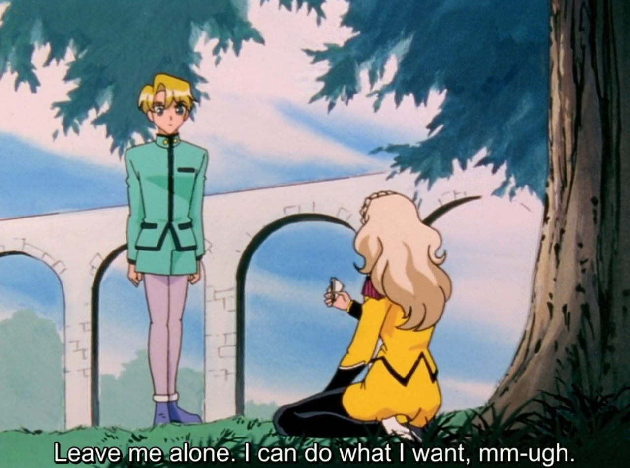 Nanami kneels on the grass under a tree, holding up a small object toward a blond boy in a green uniform who stands looking down at her. The subtitle reads “Leave me alone. I can do what I want, mm-ugh.”